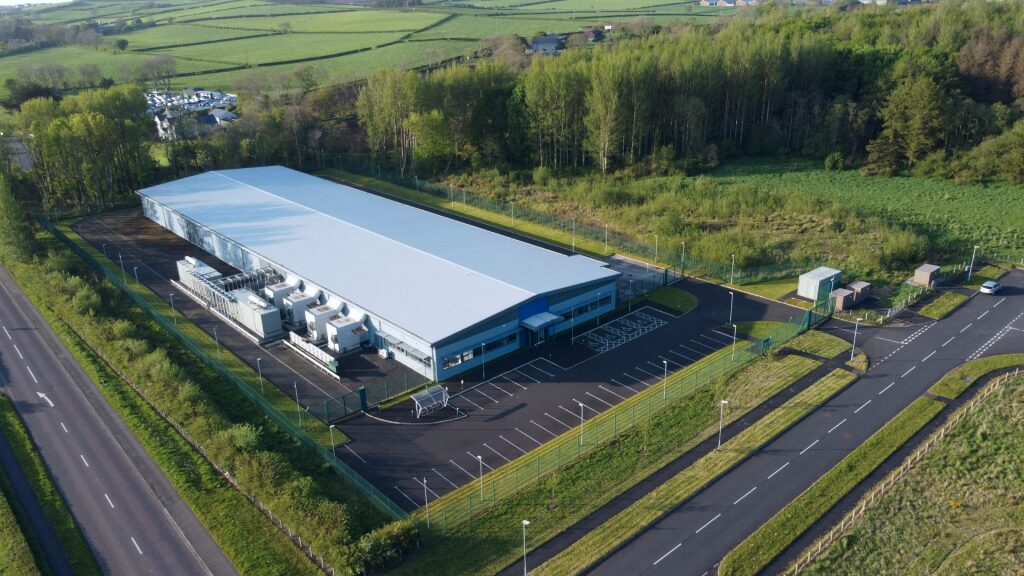 Aerial view of a single-storey datacentre with a pale grey roof and blue cladding, external plant units along one side, an empty marked car park behind perimeter fencing, woodland and green fields beyond