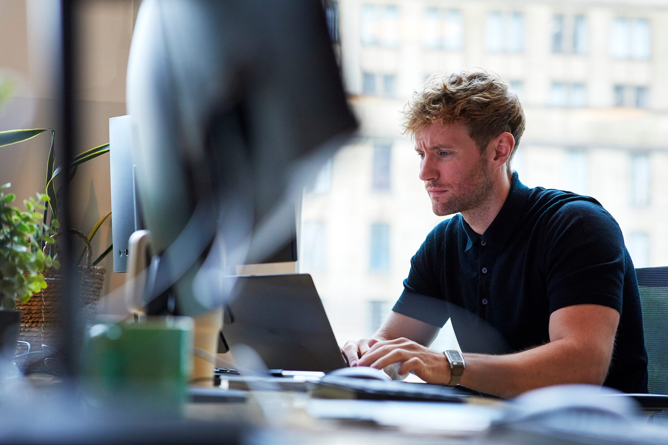 A man with curly fair hair and a short beard, in a dark navy short-sleeved polo shirt and a leather-strapped watch, typing on a laptop at a desk beside a large monitor, with a trailing green plant to his left and a city office block visible through the window behind him