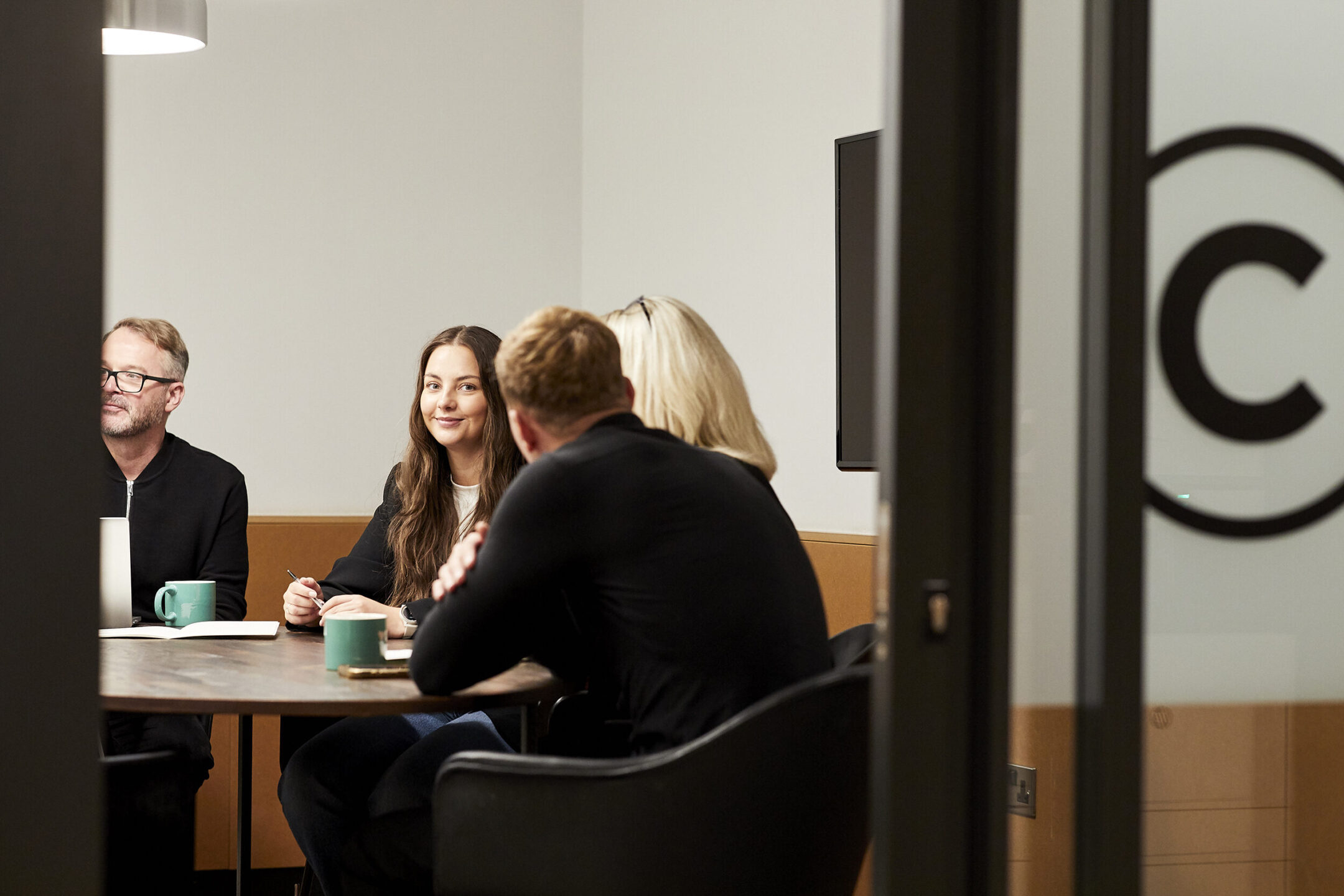 Four colleagues around a round table in a glass-walled meeting room, teal mugs and an open notebook on the table
