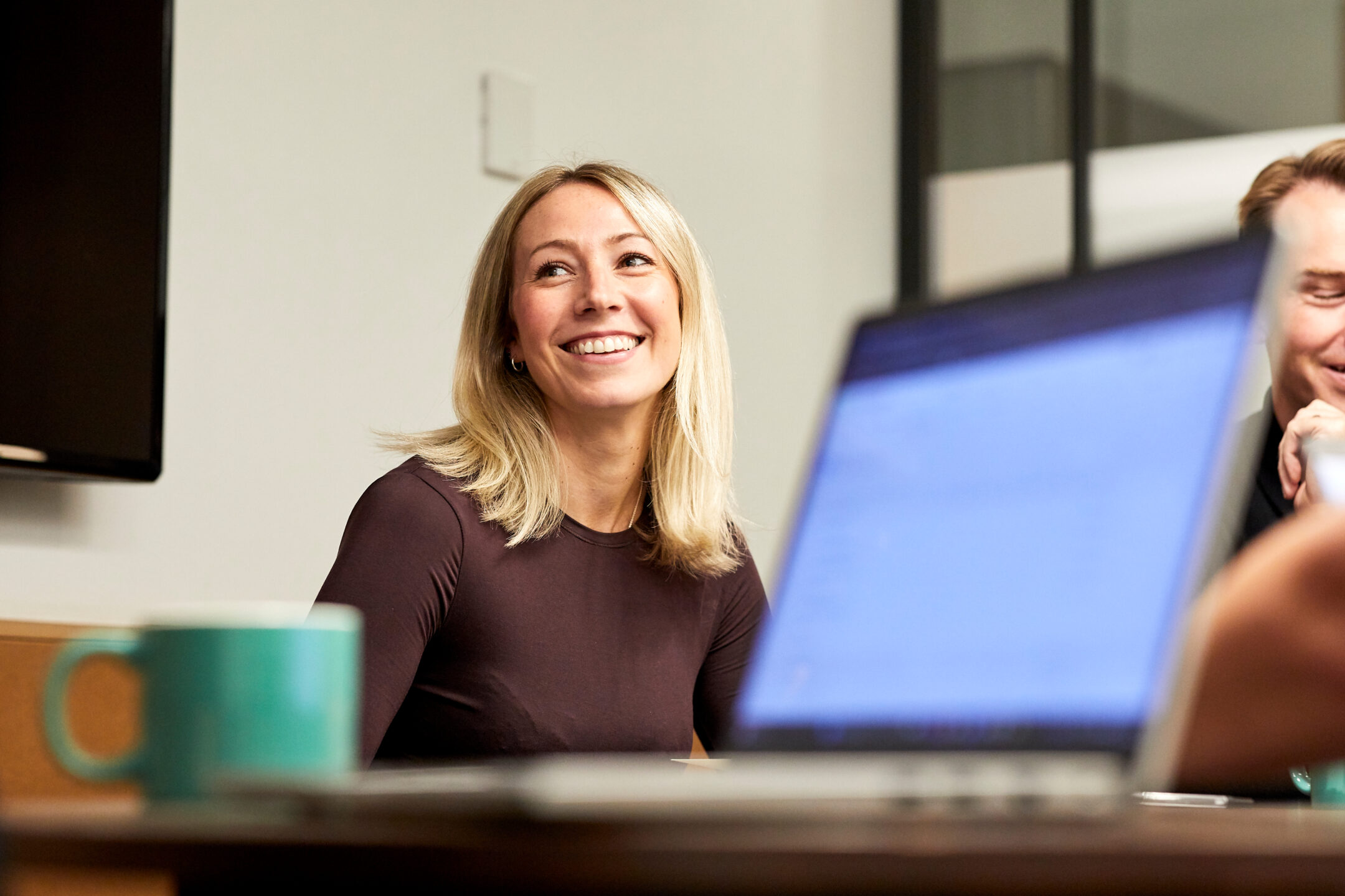 A colleague smiling while working at a laptop, a teal mug on the desk