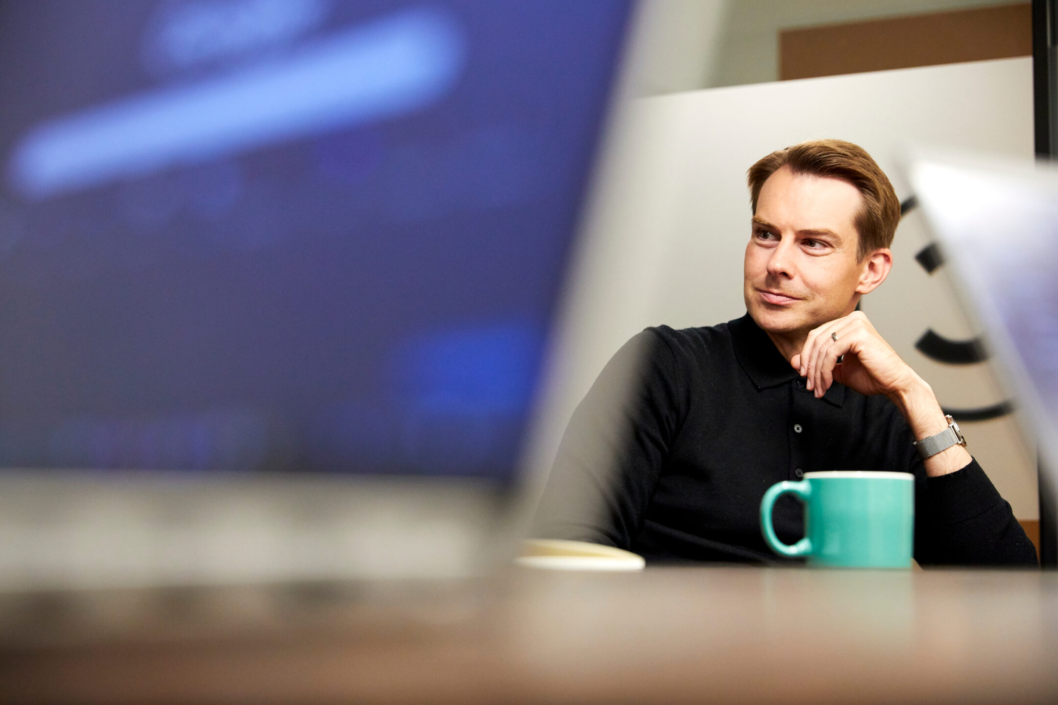 A colleague listening during a meeting, a green mug on the table beside him