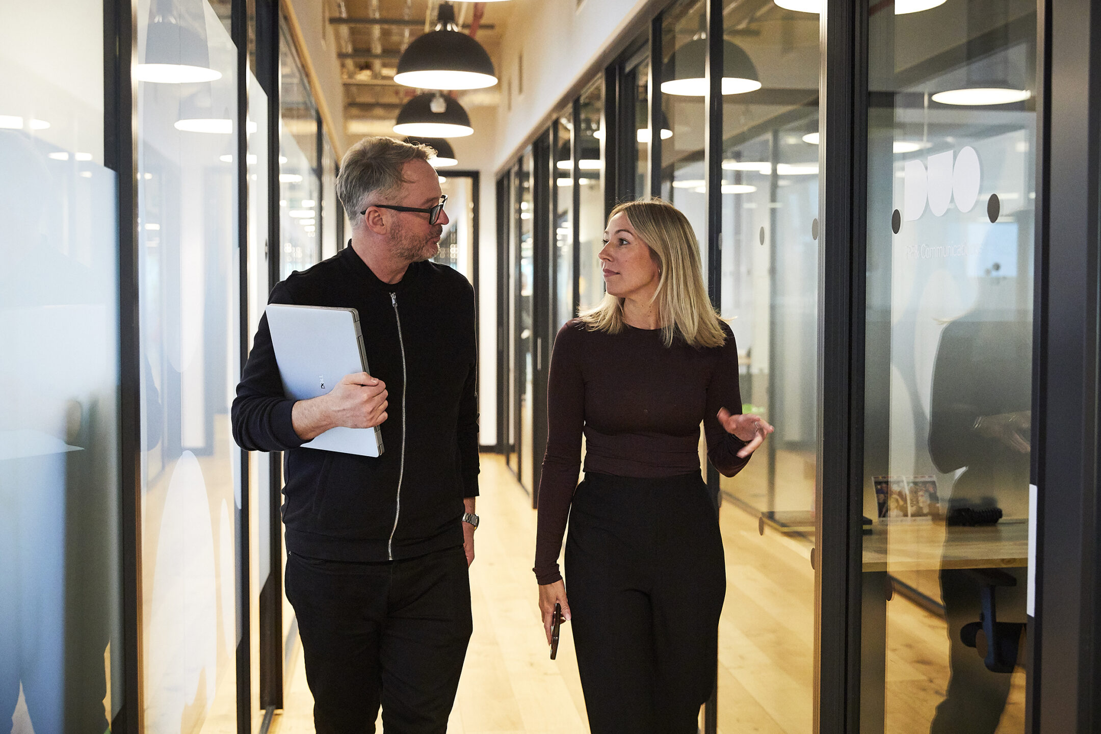 Two colleagues in conversation as they walk along a glass-walled office corridor