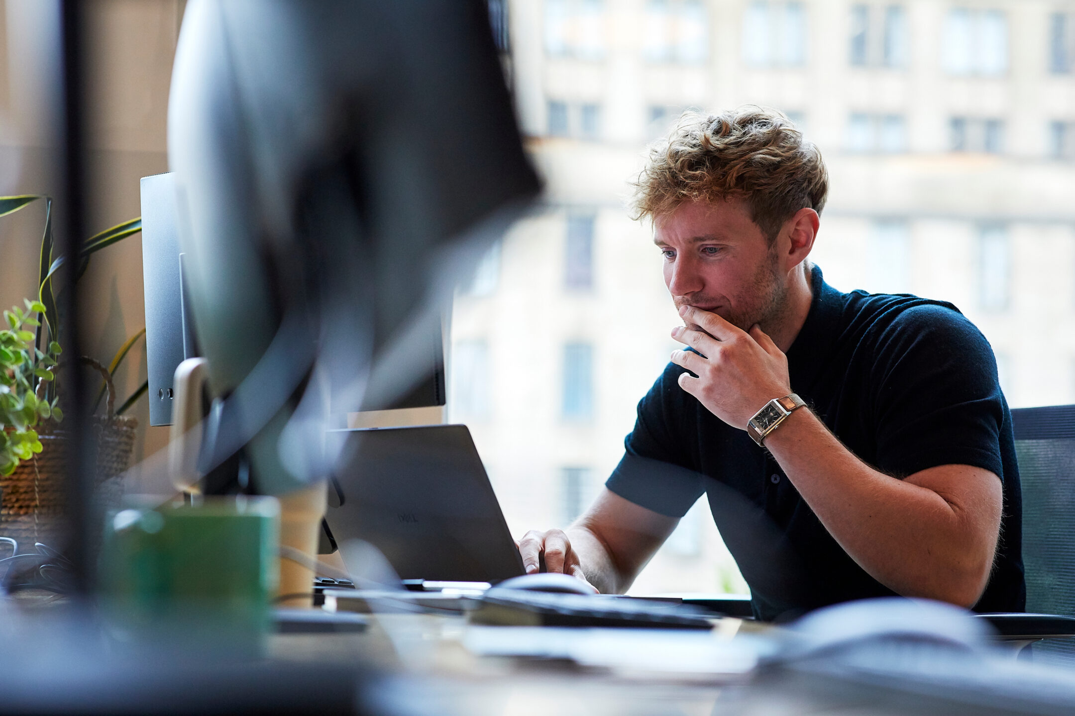 A man in a navy polo shirt at his desk, hand to his chin, looking at a laptop